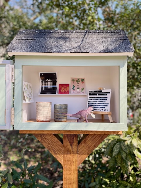 Visual Description: A Free Little Art Gallery stands in front of a large bush. The exterior of the Gallery is painted a sage color with white trim, and the interior is painted white. The roof is composed from black shingles and the structure sits on a light brown post. There are two display shelves inside the Gallery and a wooden plank floor. A small plastic dinosaur stands within; she has a tiny paint palette in hand and is holding a dark blue paintbrush. An easel positioned next to her holds a small white canvass. The following typed words are visible on the canvass: "Welcome to the Free Little Art Gallery. Take Art. Leave Art. Please do not remove the furniture or the dinosaur- she lives here and does not make for very good company when separated from her gallery!" The word "ROAR!" has been painted in purple paint onto the bottom of the canvass, seemingly by the dinosaur. Artwork has been left inside the Gallery as well. There is white ceramic cup and a blue/ green ceramic cup on the floor, and a black, green, and gold watercolor sketch of a branch is on one of the shelves. The other shelf holds three pieces: on the left is a black and white photograph of a stained-glass window, in the middle is a red and gold painted canvass, and on the right is an illustrated collection of potted plants, with the names of the plants written below each.